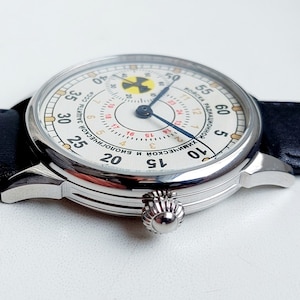 K&ouml;nnte beinhalten: Eine wei&szlig;e analoge Armbanduhr mit einem schwarzen Lederarmband. Das Zifferblatt der Uhr hat einen wei&szlig;en Hintergrund mit schwarzen Markierungen und einem blauen Sekundenzeiger. Das Zifferblatt der Uhr hat auch ein gelbes und rotes Strahlungssymbol.