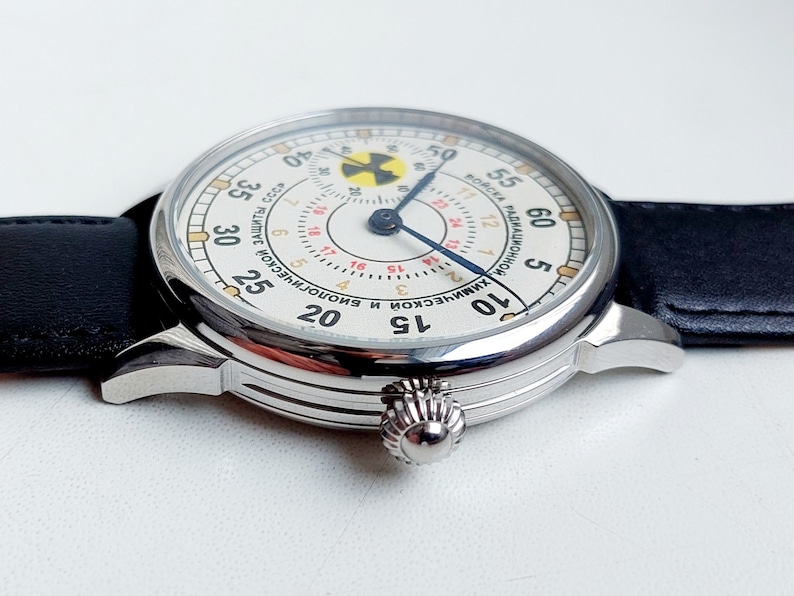 K&ouml;nnte beinhalten: Eine wei&szlig;e analoge Armbanduhr mit einem schwarzen Lederarmband. Das Zifferblatt der Uhr hat einen wei&szlig;en Hintergrund mit schwarzen Markierungen und einem blauen Sekundenzeiger. Das Zifferblatt der Uhr hat auch ein gelbes und rotes Strahlungssymbol.