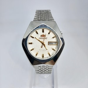 May include: A silver-toned Orient Crystal 21 Jewels wristwatch with a textured white dial, gold-colored hands and hour markers, and a day-date window displaying MON 7. The watch features a hexagonal case and a linked metal bracelet strap.