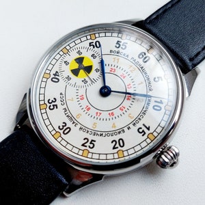 K&ouml;nnte beinhalten: Eine silberne Armbanduhr mit wei&szlig;em Zifferblatt und schwarzem Lederarmband. Das Zifferblatt der Uhr zeigt ein gelbes und schwarzes Strahlungssymbol und den Text "ВОЙСКА РАДИАЦИОННОЙ, КИМИЧЕСКОЙ И БИОЛОГИЧЕСКОЙ ЗАЩИТЫ СССР" in kyrillischer Schrift.