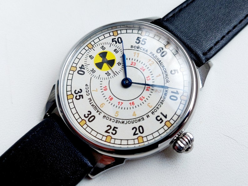 K&ouml;nnte beinhalten: Eine silberne Armbanduhr mit wei&szlig;em Zifferblatt und schwarzem Lederarmband. Das Zifferblatt der Uhr zeigt ein gelbes und schwarzes Strahlungssymbol und den Text "ВОЙСКА РАДИАЦИОННОЙ, КИМИЧЕСКОЙ И БИОЛОГИЧЕСКОЙ ЗАЩИТЫ СССР" in kyrillischer Schrift.