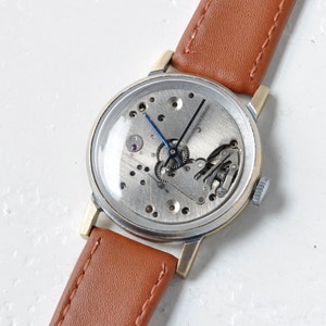 May include: A vintage wristwatch with a brown leather band and a silver face. The watch face is open, revealing the inner workings of the watch.