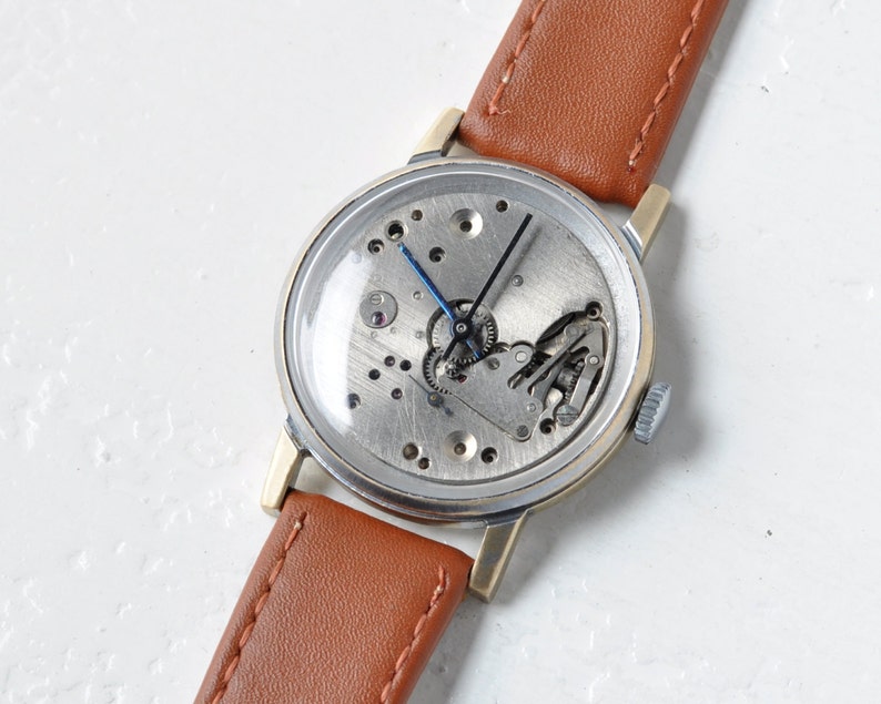 May include: A vintage wristwatch with a brown leather band and a silver face. The watch face is open, revealing the inner workings of the watch.