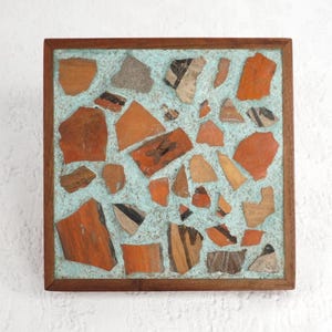 May include: A square wooden framed coaster with a turquoise blue background and a mosaic of red, brown, and black pottery shards.