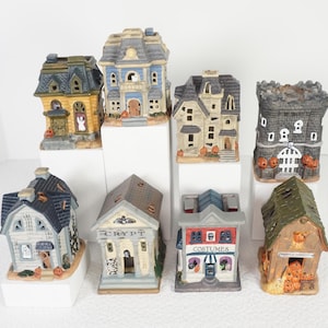 May include: A collection of eight miniature ceramic Halloween buildings, including houses, a castle, a crypt, and a costume shop. Each structure features detailed paint, including pumpkins, ghosts, and text such as CRYPT and COSTUMES on the facades.