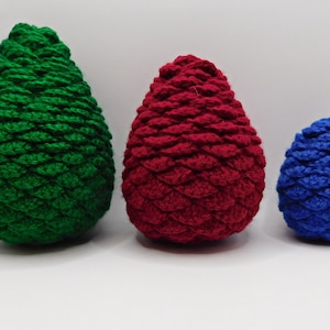 Può includere: Tre pigne all'uncinetto di diverse dimensioni. La pigna più grande è verde, la pigna di mezzo è rossa e la pigna più piccola è blu.