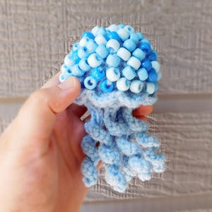 May include: A small jellyfish-shaped ornament crafted from light blue yarn and decorated with a cluster of translucent and opaque blue and white plastic pony beads on the top, featuring several dangling, spiraled crochet tentacles.