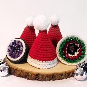 Può includere: Una composizione festosa con tre cappelli di Babbo Natale rossi e bianchi all'uncinetto con pompon bianchi, insieme a una geode all'uncinetto e un ornamento natalizio. Gli oggetti sono esposti su una fetta di legno, con ornamenti aggiuntivi.