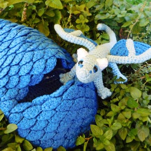 Könnte beinhalten: Ein gehäkeltes blaues Drachenei mit einer strukturierten Schuppenoptik und einem kleinen, hellblauen gehäkelten Drachen mit weißen Akzenten, der oben herauskommt. Der Drache hat dunkle Augen und kleine Flügel und ruht an der Seite des Eies.