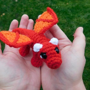 May include: A small, crocheted dragon toy with orange wings and red body. The dragon has white horns and black eyes.