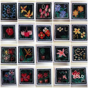 May include: A set of 26 cross-stitch embroidery pieces, each featuring a different letter of the alphabet and a floral design. The pieces are framed in black and have a white background.