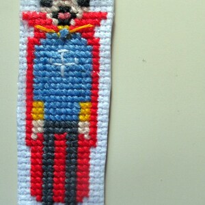 Puede incluir: Un marcapáginas hecho a mano con punto de cruz que representa un personaje pixelado con túnica azul, capa roja y pelo negro. El personaje tiene la cara bronceada y lleva zapatos negros. El marcapáginas está hecho sobre tela blanca.