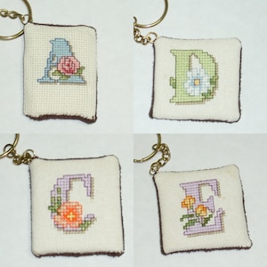 以下が含まれることがあります： A、C、D、Eの文字が刺繍されたキーホルダーが4つあります。各文字は花柄で囲まれています。キーホルダーは白で、茶色の縁取りがされています。