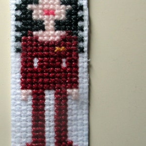 May include: Cross-stitch embroidery of a person with black hair wearing a red outfit. The person is standing with their arms at their sides.