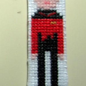 May include: A red, black, and white cross-stitch of a person wearing a red uniform with a gold emblem. The person has a bald head and a pink face.