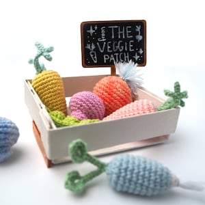 May include: A white wooden crate filled with crocheted vegetables, including carrots in yellow, orange, and pink, and a mushroom in pink. A sign above the crate reads "from THE VEGGIE PATCH".