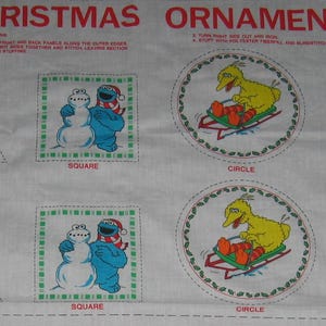 May include: A white fabric panel with the words "Christmas Ornaments" printed in red. The panel features nine different Sesame Street character designs, including Big Bird, Elmo, Cookie Monster, and Oscar the Grouch. Each design is labeled with a shape: circle, triangle, or square.