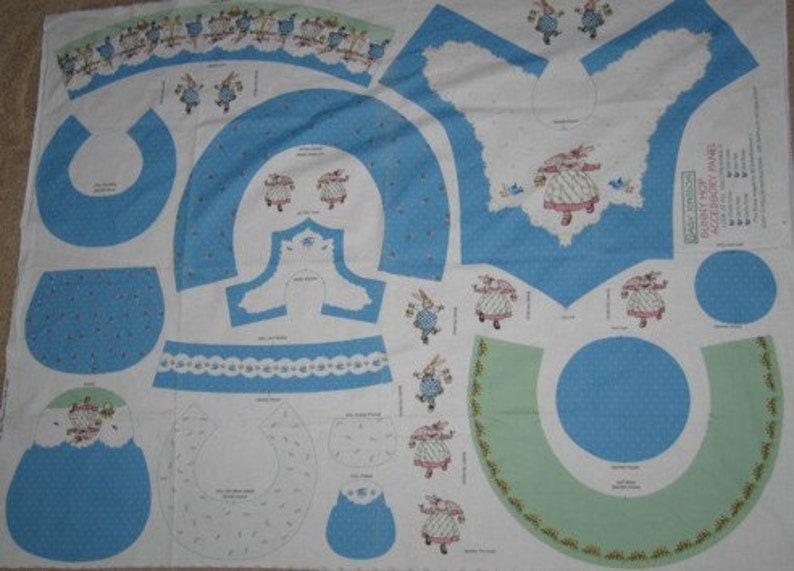 May include: A fabric panel with a blue and white pattern featuring a bunny rabbit wearing a dress and holding a bird. The panel includes multiple pieces for sewing a bunny rabbit costume. The text on the panel reads "Bunny Hat Panel - Accessories for the Bunny Hat."