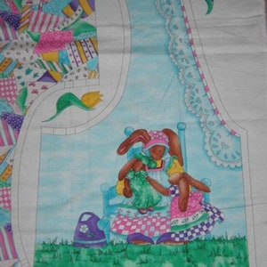 May include: A fabric panel with a blue background featuring a mother bunny and a baby bunny sitting on a chair. The mother bunny is wearing a pink and white dress and a pink hat. The baby bunny is wearing a green dress. The fabric panel is surrounded by a green border and a white border.