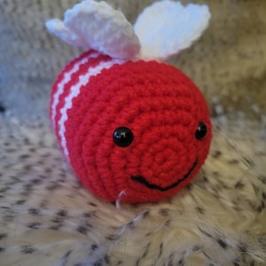May include: A crocheted red and white bee with black eyes and a smiling mouth. The bee has white wings.