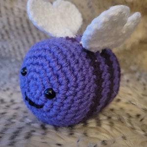 May include: A crocheted purple and white bee plush toy with black eyes and a smiling mouth. The bee has white wings and is sitting on a fuzzy white surface.