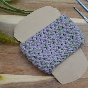 May include: A rectangular piece of crocheted fabric featuring a variegated pattern of light purple and sage green yarn. The textile is wrapped around a plain, light brown cardboard display card, showcasing the textured, interlocking stitch pattern.