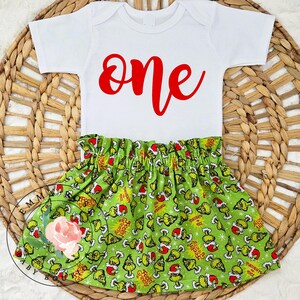 May include: A white short-sleeved bodysuit with the word one printed in red script lettering, paired with a green skirt featuring an all-over pattern of cartoon characters wearing red Santa hats and the text Feelin Grinchy.