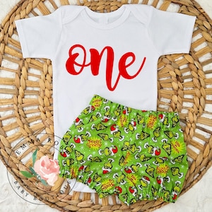 May include: A white short-sleeved bodysuit with the word one printed in red script lettering, paired with green bloomers featuring an all-over print of cartoon characters wearing Santa hats and the text Feelin Grinchy.