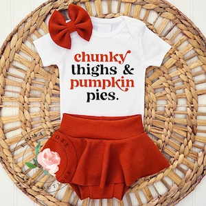 May include: A white short-sleeved bodysuit with the text chunky thighs & pumpkin pies in orange and black lettering, paired with a rust-colored textured bloomer skirt and a matching rust-colored hair bow with a ribbed fabric texture.