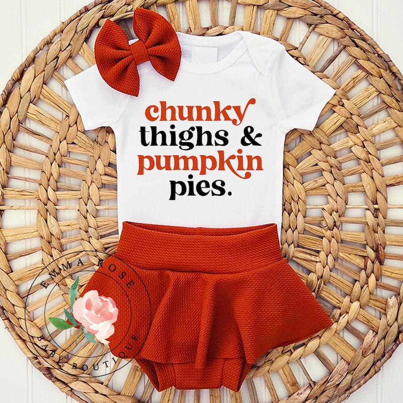 May include: A white short-sleeved bodysuit with the text chunky thighs & pumpkin pies in orange and black lettering, paired with a rust-colored textured bloomer skirt and a matching rust-colored hair bow with a ribbed fabric texture.