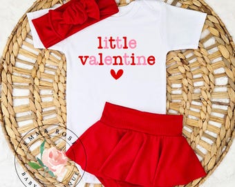 May include: A white baby bodysuit with the words "little valentine" in red and pink, and a red heart. The outfit includes a red headband and a red skirt. The items are arranged on a woven basket.