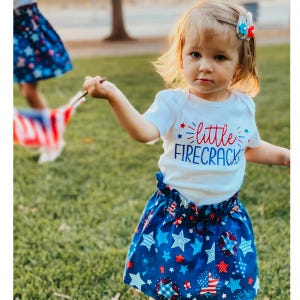 Op de afbeelding: Een jong kind draagt een witte romper met de tekst "little FIRECRACKER" in rood en blauw. Het kind draagt ook een blauwe rok met een patriottisch sterrenpatroon. Het kind houdt een kleine Amerikaanse vlag vast.
