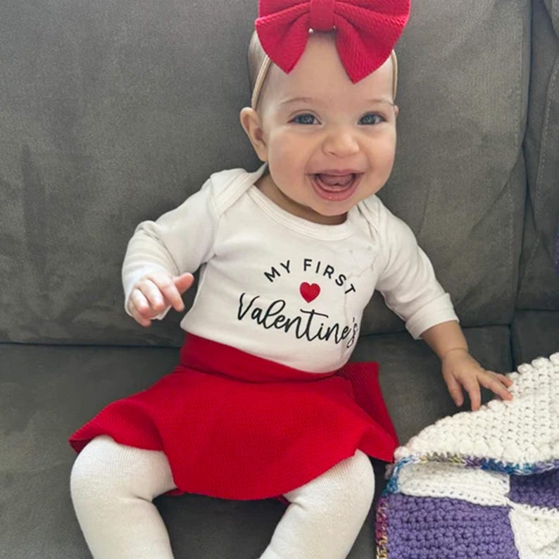May include: A long-sleeved white bodysuit with the text My First Valentine's and a small red heart printed on the front. The outfit includes a bright red skirt, white leggings, and a large red bow headband worn on the head.
