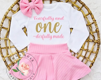 May include: A white long-sleeve bodysuit with the pink and gold glitter text Fearfully and one-derfully made, paired with a pink ribbed skirt and a matching pink ribbed hair bow. The set is displayed on a woven circular basket.