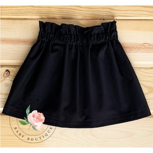 May include: A solid black skirt featuring a high-waisted paper bag style with a gathered, ruffled elastic waistband. The skirt has a flared, A-line silhouette and includes a circular logo with a pink rose and the text Emma Rose Baby Boutique.