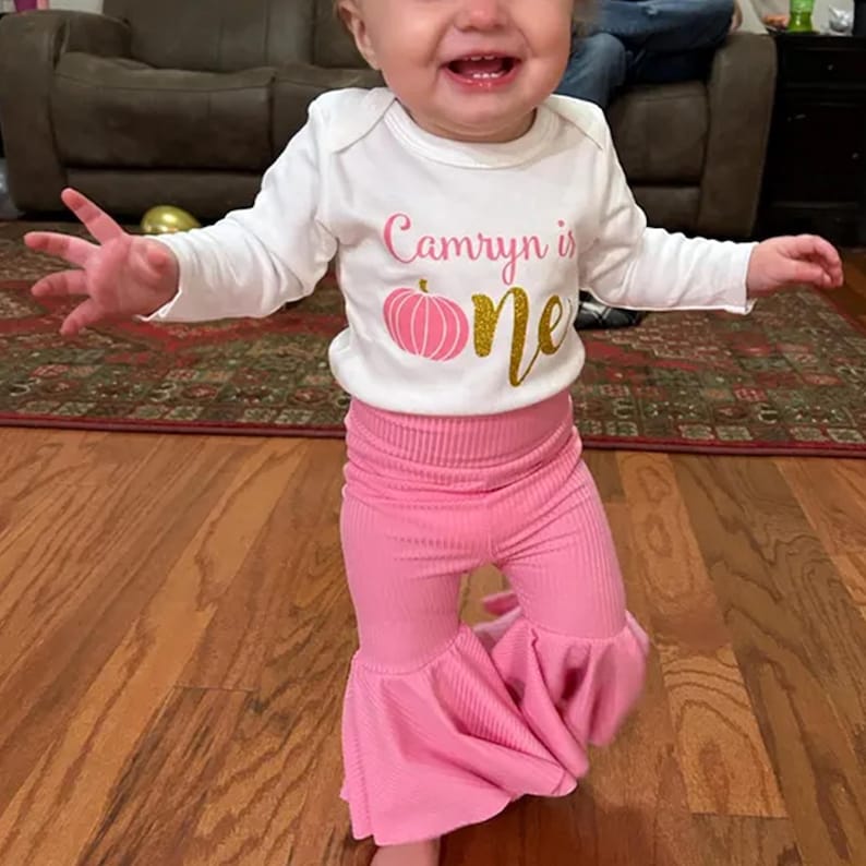 Pu&ograve; includere: Un body bianco a maniche lunghe con la scritta "Camryn is" in rosa, una grafica a forma di zucca rosa e la parola "one" in glitter oro. Il body &egrave; abbinato a pantaloni a zampa a coste rosa tinta unita con vita alta elastica.