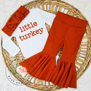 Pu&ograve; includere: Un body bianco a maniche lunghe con la scritta little turkey in arancione, abbinato a pantaloni a zampa a coste color ruggine e una fascia per capelli coordinata a coste color ruggine con un grande fiocco. Il set &egrave; presentato come completo.