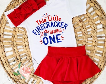 May include: A white short-sleeved bodysuit with the text This Little FIRECRACKER is TURNING ONE in red and blue, paired with a red ribbed headband featuring a bow and a matching red ribbed skirted bloomer with a flared hemline design.