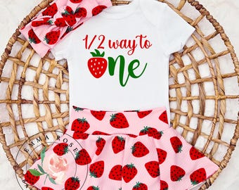 Può includere: Un completo per neonati con un body bianco con la scritta "1/2 way to one" e una fragola. Il set include una fascia e pantaloncini rosa abbinati, entrambi decorati con motivi di fragole rosse. L'outfit è presentato su un cestino intrecciato.