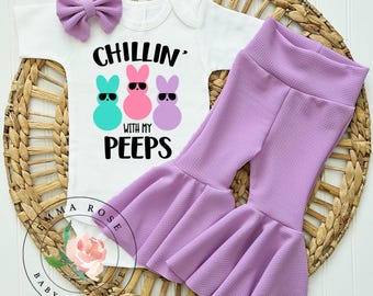 May include: A white baby bodysuit with the text "CHILLIN' WITH MY PEEPS" and three cartoon bunnies wearing sunglasses. The outfit includes lavender bell-bottom pants and a matching bow. The set is displayed on a woven basket.