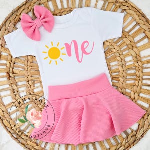 May include: A white short-sleeved bodysuit featuring a yellow sun graphic and the pink text "one", paired with a pink textured skirt and a matching pink hair bow. The set includes a circular logo with a floral illustration and the text "EMMA ROSE BABY BOUTIQUE".