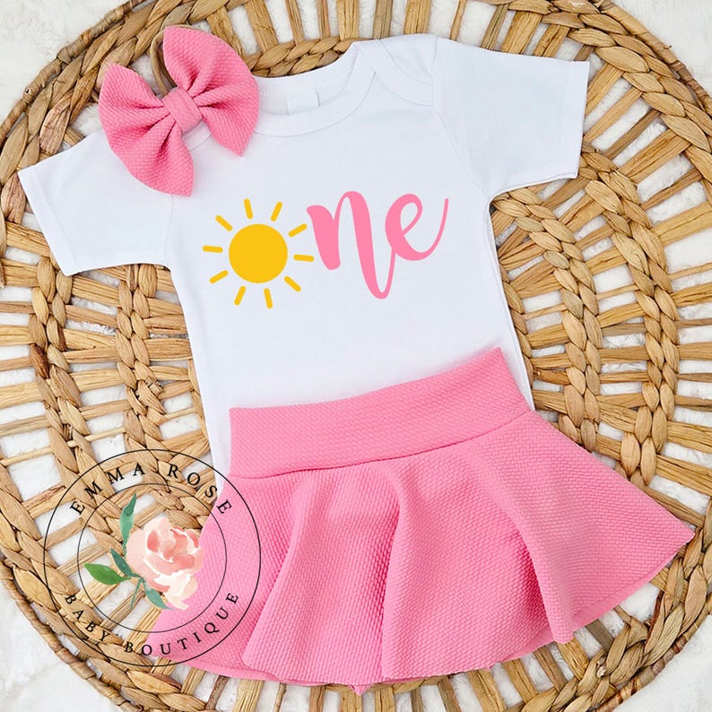 May include: A white short-sleeved bodysuit featuring a yellow sun graphic and the pink text "one", paired with a pink textured skirt and a matching pink hair bow. The set includes a circular logo with a floral illustration and the text "EMMA ROSE BABY BOUTIQUE".