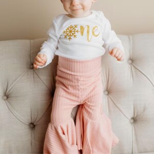Pu&ograve; includere: Un body bianco a maniche lunghe con una grafica a fiocco di neve glitterata oro e la parola one in corsivo glitterato oro, abbinato a pantaloni a zampa d'elefante a coste rosa chiaro con vita alta e linea svasata.