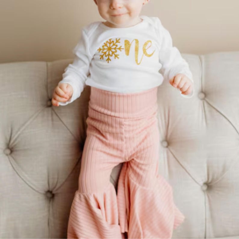 Pu&ograve; includere: Un body bianco a maniche lunghe con una grafica a fiocco di neve glitterata oro e la parola one in corsivo glitterato oro, abbinato a pantaloni a zampa d'elefante a coste rosa chiaro con vita alta e linea svasata.