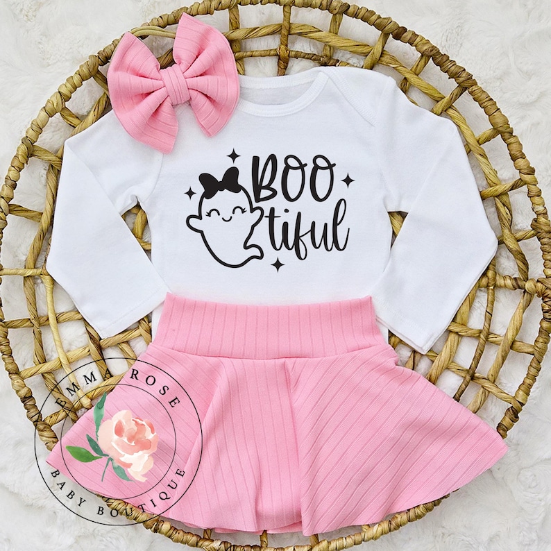 May include: A white long-sleeve bodysuit with a black graphic of a ghost wearing a bow and the text BOO tiful, paired with a light pink ribbed skirt and a matching pink ribbed hair bow. The set features a clean design with simple lines.