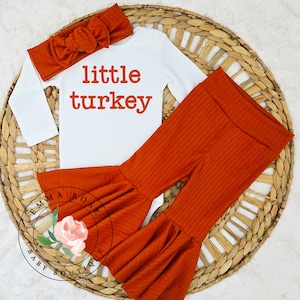 Pu&ograve; includere: Un body bianco a maniche lunghe con la scritta little turkey in arancione, abbinato a pantaloni a zampa a coste arancioni e una fascia per capelli a coste coordinata con un grande dettaglio a fiocco sulla parte superiore.