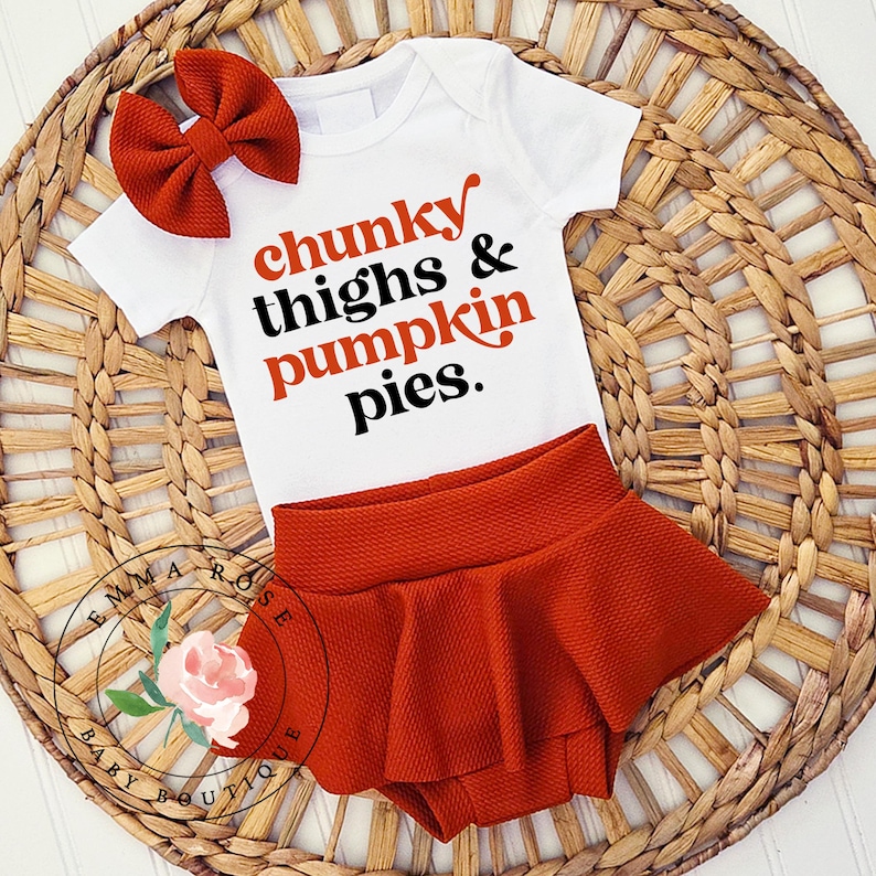 May include: A white short-sleeved baby bodysuit with the text chunky thighs & pumpkin pies. in orange and black lettering, paired with a matching burnt orange textured bow headband and a matching burnt orange textured ruffled bloomer skirt.