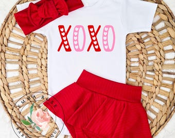 May include: A white short-sleeved bodysuit with the text XOXO in red and pink, featuring heart patterns within the letters. The set includes a red ribbed headband with a bow and a matching red ribbed skirted bloomer with a flared hemline.