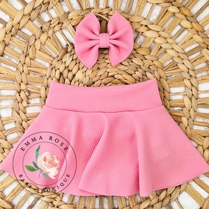 May include: A solid pink skirt with a textured fabric finish and a matching pink bow hair accessory. The skirt features a flared silhouette with a wide waistband. A circular logo with the text Emma Rose Baby Boutique and a floral graphic is visible.