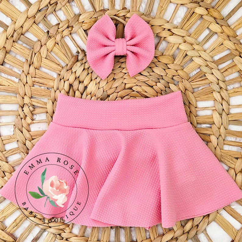 May include: A solid pink skirt with a textured fabric finish and a matching pink bow hair accessory. The skirt features a flared silhouette with a wide waistband. A circular logo with the text Emma Rose Baby Boutique and a floral graphic is visible.